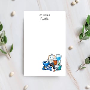 May include: A white rectangular notepad with the text FROM THE DESK OF Nicole printed at the top. The bottom right corner features a colourful illustration of medical supplies including a clipboard, mortar and pestle, pills, IV bag, and syringe.
