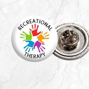 May include: A white button with a colourful handprint design and the text "Recreational Therapy". The button is attached to a metal pin back.