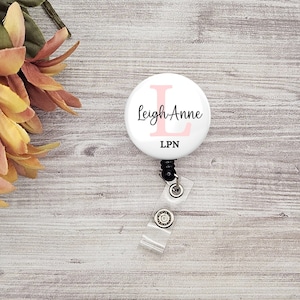 May include: A white badge reel featuring the name "Leigh Anne" in black script and a pink "L" initial. The letters "LPN" are printed in black below the name. The reel has a clear plastic strap and a black retractable cord.