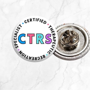 May include: A white button with the text "CTRS" in colorful letters. The text around the button reads "Certified Therapeutic Recreation Specialist". The button is attached to a silver pin back.