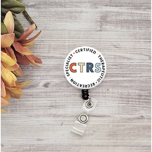 May include: A white CTRS badge reel with the text "SPECIALIST CERTIFIED THERAPEUTIC RECREATION". The letters CTRS are in orange, teal, and tan. The badge reel has a clear plastic strap and a black retractable cord.
