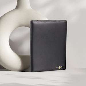 May include: A black leather portfolio with gold-colored initials "jw" in a script font. The portfolio is rectangular and stands upright. A cream-colored, abstract ceramic vase is in the background.