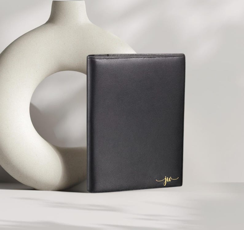 May include: A black leather portfolio with gold-colored initials "jw" in a script font. The portfolio is rectangular and stands upright. A cream-colored, abstract ceramic vase is in the background.