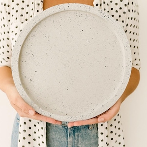 Puede incluir: Una bandeja redonda de hormigón moteado en color gris claro. La bandeja la sostiene una persona que lleva una camisa blanca con lunares negros y vaqueros azules. La bandeja tiene aproximadamente 30 cm de diámetro.