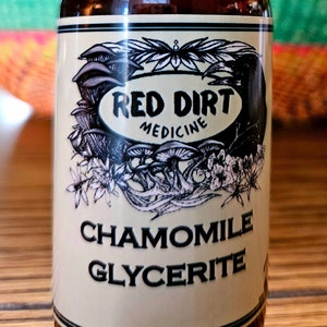 May include: A brown glass bottle with a dropper top and a white label. The label has black text that reads "Red Dirt Medicine" and "Chamomile Glycerite". The label also features a black and white illustration of flowers and mushrooms.