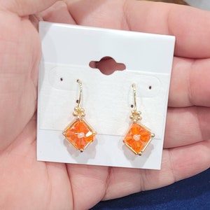 May include: A pair of gold earrings with orange crystal flower accents. The earrings are on a white card.