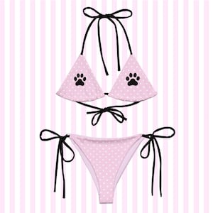 May include: A two-piece bikini set featuring a light pink fabric with a white polka dot pattern. The triangle top and bottom are accented with black ties and a black paw print graphic on each side of the top. The set includes adjustable strings.