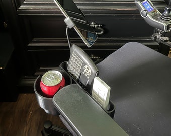 May include: A black wheelchair with a variety of accessories, including a phone holder, a keyboard, a remote control, and a cup holder. The phone holder is attached to a flexible arm and is positioned above the keyboard. The keyboard is in a black case and is positioned next to the remote control. The cup holder is on the armrest of the wheelchair and is holding a red can.