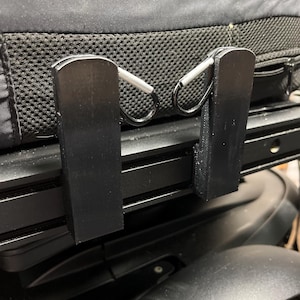 May include: Two black plastic hooks attached to the back of a black chair. The hooks have silver clips.