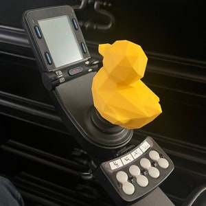 May include: A yellow, geometrically-shaped rubber ducky sits atop a black joystick with a small screen and buttons. The joystick is mounted on a black arm.
