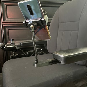 May include: A black phone holder with a flexible arm attached to a black chair. The phone holder is holding a black smartphone.