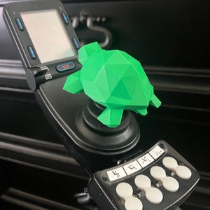 May include: A bright green, geometric turtle figurine sits atop a black joystick control panel. The panel features a digital display and several white buttons with icons. The turtle is a 3D-printed design.