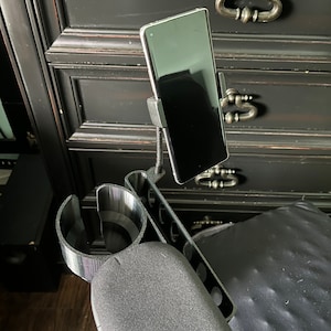 May include: A black phone holder with a cup holder attached to a black armrest. The phone holder is holding a black smartphone.