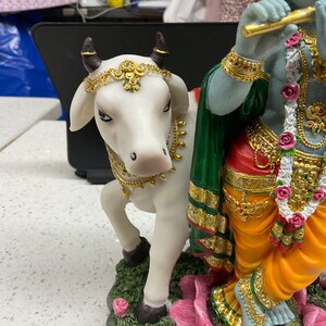K&ouml;nnte beinhalten: Eine wei&szlig;e Kuhfigur mit goldenen Akzenten steht neben einer blauen und gr&uuml;nen Figur von Krishna, der Fl&ouml;te spielt. Die Krishna-Figur tr&auml;gt einen gelben Dhoti und eine rot-gr&uuml;ne Sch&auml;rpe. Die Kuh tr&auml;gt eine goldene Halskette und ein goldenes Stirnband mit gr&uuml;nen und roten Juwelen. Die Figuren stehen auf einer rosa Lotusbl&uuml;te.