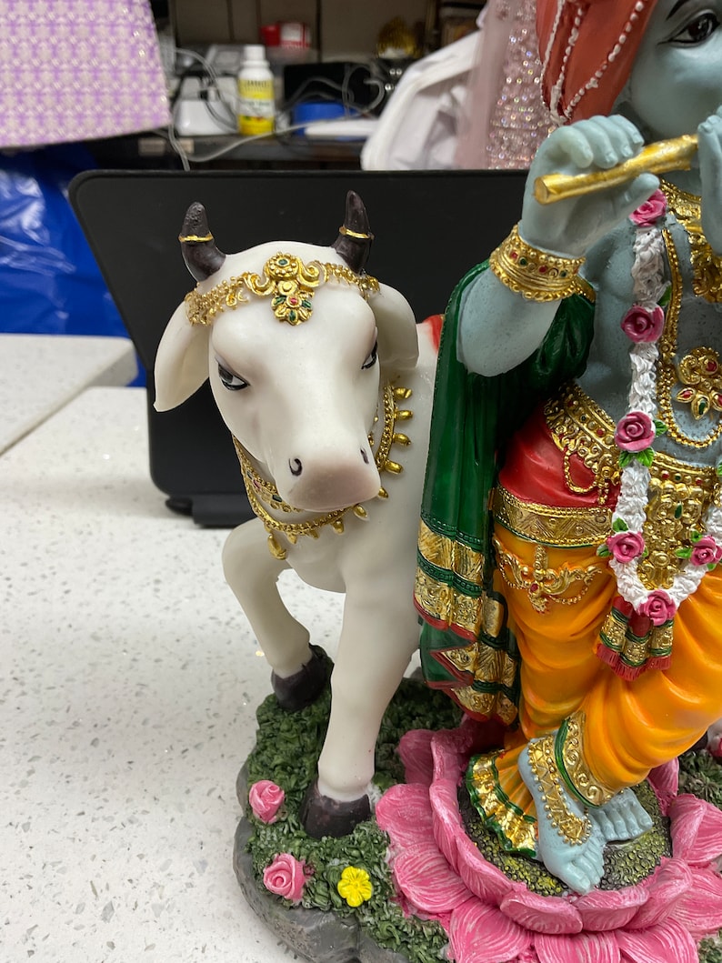 K&ouml;nnte beinhalten: Eine wei&szlig;e Kuhfigur mit goldenen Akzenten steht neben einer blauen und gr&uuml;nen Figur von Krishna, der Fl&ouml;te spielt. Die Krishna-Figur tr&auml;gt einen gelben Dhoti und eine rot-gr&uuml;ne Sch&auml;rpe. Die Kuh tr&auml;gt eine goldene Halskette und ein goldenes Stirnband mit gr&uuml;nen und roten Juwelen. Die Figuren stehen auf einer rosa Lotusbl&uuml;te.