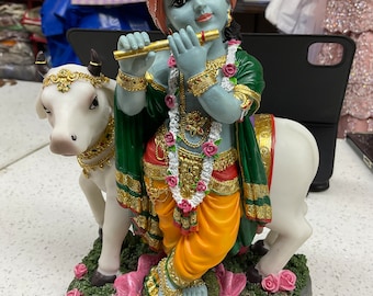 May include: A blue and green figurine of Krishna playing a flute, standing on a pink lotus flower and riding a white cow. The figurine is decorated with gold accents and flowers.