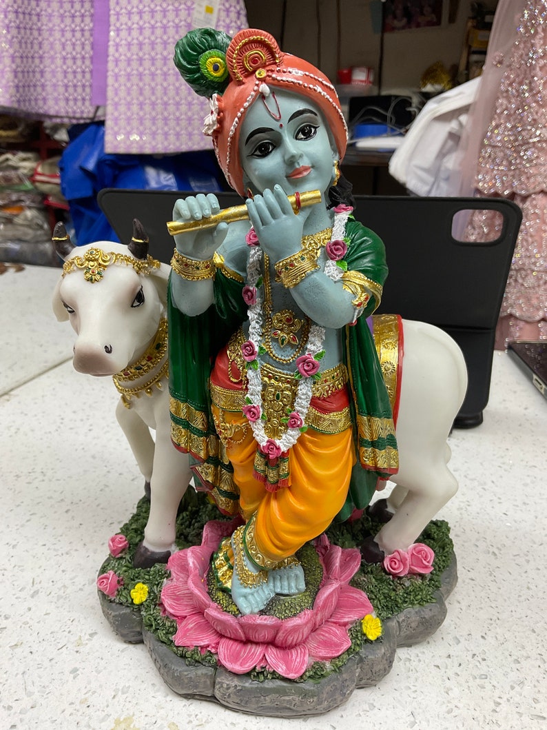 K&ouml;nnte beinhalten: Eine blaue und gr&uuml;ne Figur von Krishna, der Fl&ouml;te spielt, auf einer rosa Lotusbl&uuml;te steht und auf einem wei&szlig;en Kuh reitet. Die Figur ist mit goldenen Akzenten und Blumen verziert.