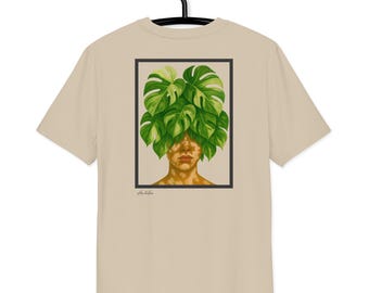 Puede incluir: Una camiseta beige colgada de una percha negra. La parte posterior de la camiseta presenta una ilustración enmarcada de la cara de una persona con hojas verdes de monstera que cubren la cabeza. La obra de arte tiene un tema botánico natural.