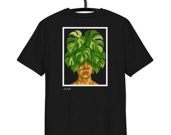 Puede incluir: Camiseta negra con una ilustración enmarcada de una persona con hojas verdes de monstera cubriendo su cabeza. La obra de arte tiene un borde blanco y está sobre un fondo negro. La camiseta está colgada de una percha negra.