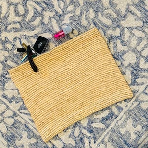 May include: A woven straw clutch bag with a zipper closure. The bag is open and has a few items inside, including a key chain, a lip gloss, and a small silver object.