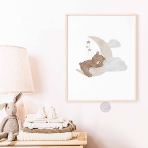 May include: A watercolor illustration of a brown bear sleeping on a crescent moon with stars and clouds. The illustration is in a light gray and brown color palette.