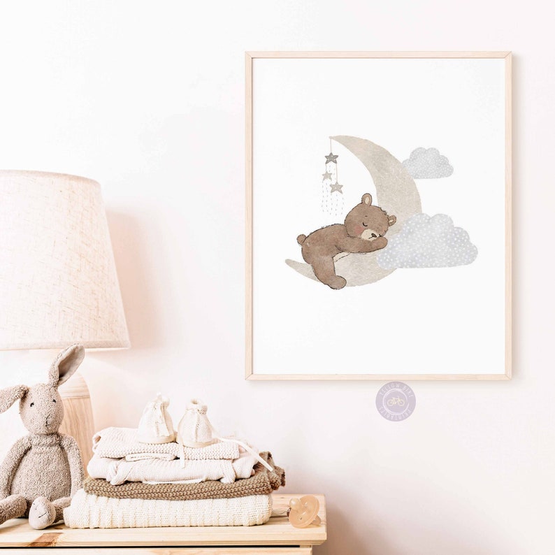 May include: A watercolor illustration of a brown bear sleeping on a crescent moon with stars and clouds. The illustration is in a light gray and brown color palette.
