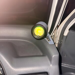 May include: A car dashboard detail showing a digital gauge with a yellow display. The gauge is mounted on a grey bracket and set against a grey dashboard. The display reads "0.0".