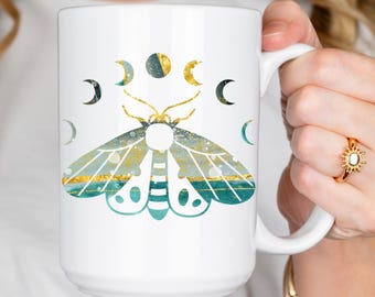 May include: White ceramic mug featuring a moth design with teal, gold, and white accents. The moth is surrounded by crescent moon phases. The mug has a large handle for easy gripping and is suitable for hot or cold beverages.