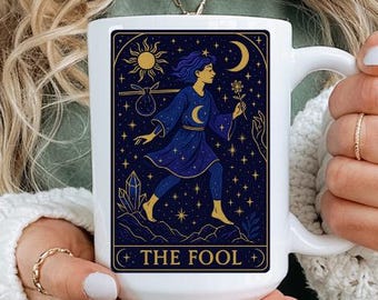 May include: White ceramic mug featuring a tarot card design with a figure in a blue robe, a crescent moon, and the text "THE FOOL." The design is framed by a gold border. The mug is held by a person wearing a green shirt and a white sweater.