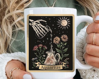 May include: White ceramic mug featuring an illustration of a skeleton hand pouring water into a decorative pitcher, with flowers and the sun. The word "Aquarius" is printed on the mug. The mug is held by a person.