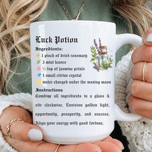 May include: White ceramic mug with the text "Luck Potion" and a list of ingredients and instructions. The mug features a floral illustration and is held by a person wearing a green shirt and a white sweater.