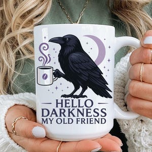 May include: White ceramic mug featuring a black raven illustration with a crescent moon and coffee cup design. The mug has the text "Hello Darkness My Old Friend" in black font. The mug has a white handle.