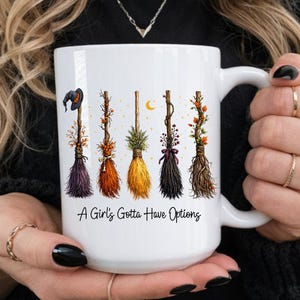 Puede incluir: Taza de cer&aacute;mica blanca con seis dise&ntilde;os decorativos de escobas en varios colores. Las escobas est&aacute;n adornadas con detalles florales y oto&ntilde;ales. La taza tiene el texto "A Girl's Gotta Have Options" en negro.