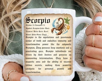 May include: White ceramic mug featuring the word "Scorpio" in a vintage style font, with a scorpion illustration and descriptive text. The mug is held by a person wearing a green shirt and a white sweater.