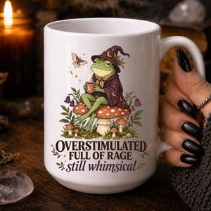 Puede incluir: Una taza de cerámica blanca con el gráfico de una rana verde con sombrero y túnica de mago de color morado, sentada sobre una seta roja con puntos blancos. El texto dice: OVERSTIMULATED FULL OF RAGE still whimsical. Una pequeña polilla y flores rodean a la rana.