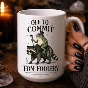 May include: White ceramic mug with the text "OFF TO COMMIT TOM FOOLERY" and a whimsical illustration of a frog riding a raccoon. The frog wears a top hat and holds a cane. The phrase "with malicious whimsy" is printed at the bottom.