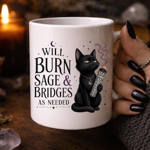 Peut inclure: Mug en c&eacute;ramique blanche avec un chat noir tenant un fagot de sauge et le texte "WILL BURN SAGE & BRIDGES AS NEEDED". Le chat porte un collier croissant de lune. La tasse est tenue par une personne avec du vernis &agrave; ongles noir.