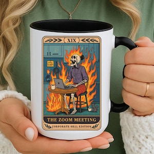 Könnte beinhalten: Weiße Keramik-Tasse mit schwarzem Innenraum und Griff. Die Tasse zeigt ein Tarotkarten-Design mit einem brennenden Skelett an einem Tisch mit einem Laptop. Der Text lautet "THE ZOOM MEETING CORPORATE HELL EDITION."