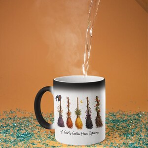 Puede incluir: Una taza m&aacute;gica que cambia de color con asa y borde negros. La taza blanca presenta un dise&ntilde;o de seis coloridas escobas de bruja y el texto "A Girl's Gotta Have Options". Se vierte agua en la taza.