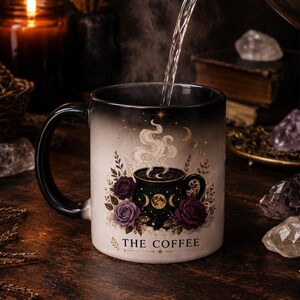 Op de afbeelding: Een zwart-witte koffiemok met een bloemen- en hemels ontwerp. De mok heeft paarse rozen, halve manen en de tekst "THE COFFEE". Er wordt heet water in de mok gegoten, waardoor stoom ontstaat.