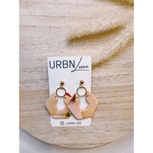 May include: A pair of peach-colored geometric earrings with gold hoops and a swirl pattern. The earrings are displayed on a white card with the text "URBN/luxe" and "@_URBNLUXE".