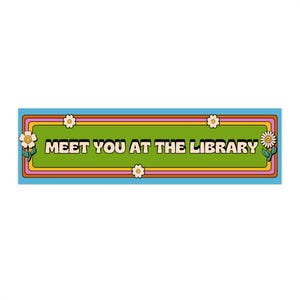 Könnte beinhalten: Ein rechteckiges Schild mit blauem Rand und grünem Zentrum. Auf dem Schild steht "MEET YOU AT THE LIBRARY" in weißer Schrift. Das Schild ist mit Blumen und einem Retro-Farbschema verziert.