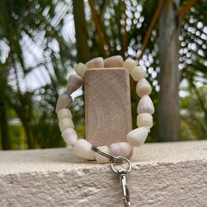 May include: A bracelet made of light-colored, irregular-shaped beads, with a silver-colored clasp. The bracelet is looped around a small, rectangular, light-brown block. The background is blurred, with green foliage.