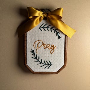 May include: An octagonal wooden embroidery hoop frame containing white fabric with a small cross pattern. The word Pray is embroidered in gold thread in the center, flanked by two dark green embroidered leafy branches. A gold satin ribbon bow is attached.
