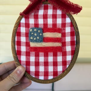 May include: A round, wooden-framed decoration featuring a red and white gingham pattern. The center displays a felted American flag with a blue field of stars and red and white stripes. A red ribbon bow adorns the top.