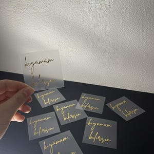 May include: A collection of square, transparent stickers with gold script. The stickers are scattered on a black surface, with one held by a hand. The text on the stickers reads "kıyamam bilirsin".