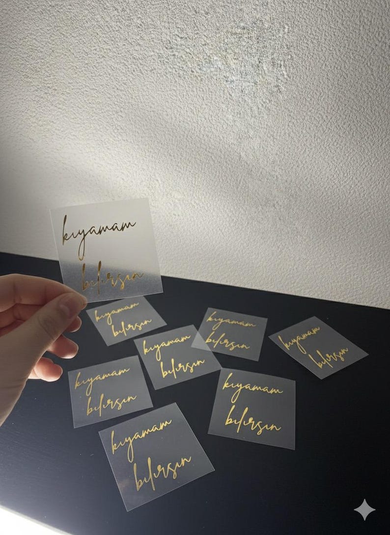 May include: A collection of square, transparent stickers with gold script. The stickers are scattered on a black surface, with one held by a hand. The text on the stickers reads "kıyamam bilirsin".
