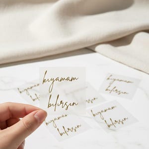 May include: Clear square stickers with gold script text that reads "kıyamam bilirsin". The stickers are held by a hand, with several others scattered on a white marble surface. A beige fabric is in the background.