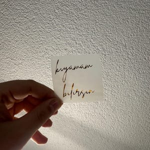 May include: A small, square white sticker with gold script text. The text reads "kıyamam" above "bilirsin". The sticker is held against a textured, off-white wall.