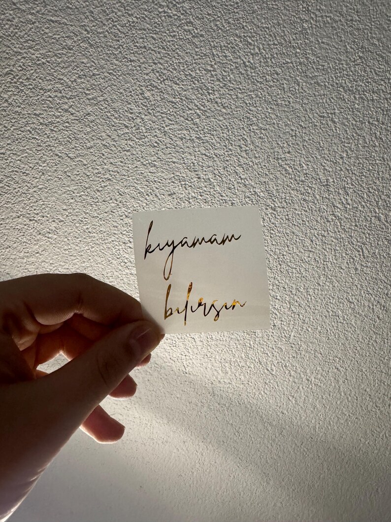 May include: A small, square white sticker with gold script text. The text reads "kıyamam" above "bilirsin". The sticker is held against a textured, off-white wall.